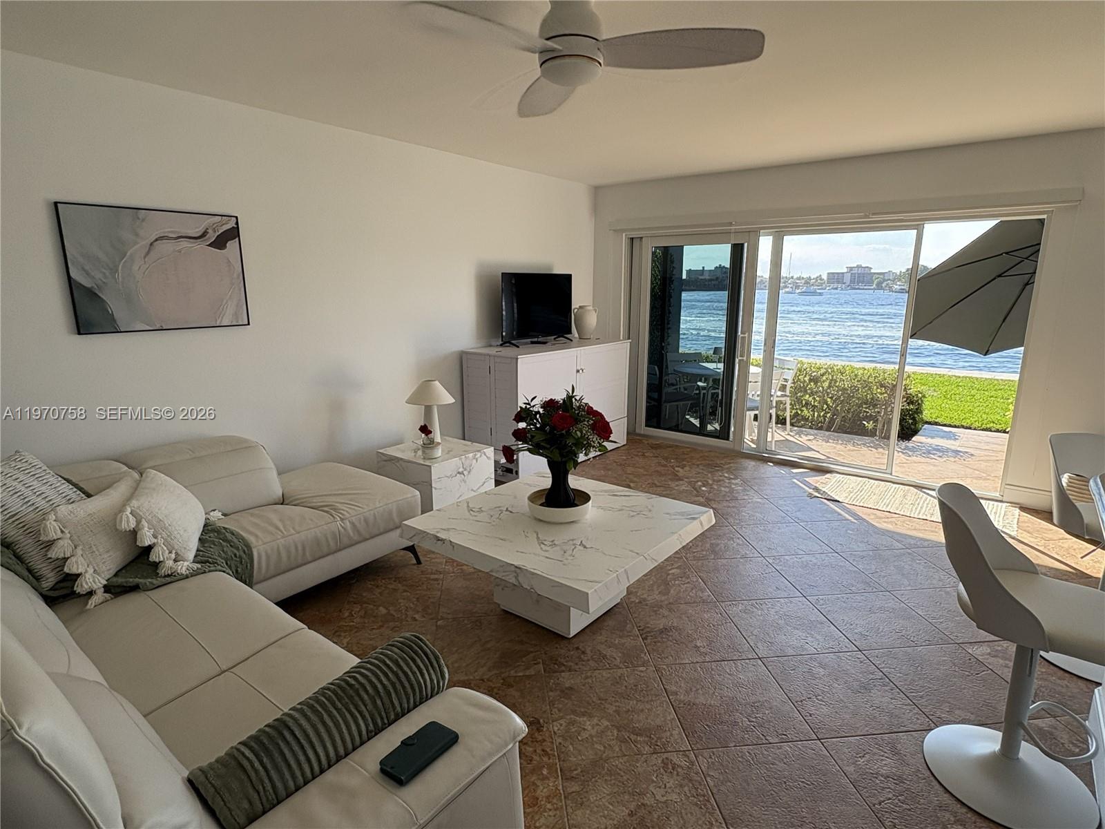 A living room features a white sectional sofa, marble coffee table, and sliding doors overlooking water — 1201 S Riverside Dr # 107, Pompano Beach FL 33062, POMPANO BEACH, FL