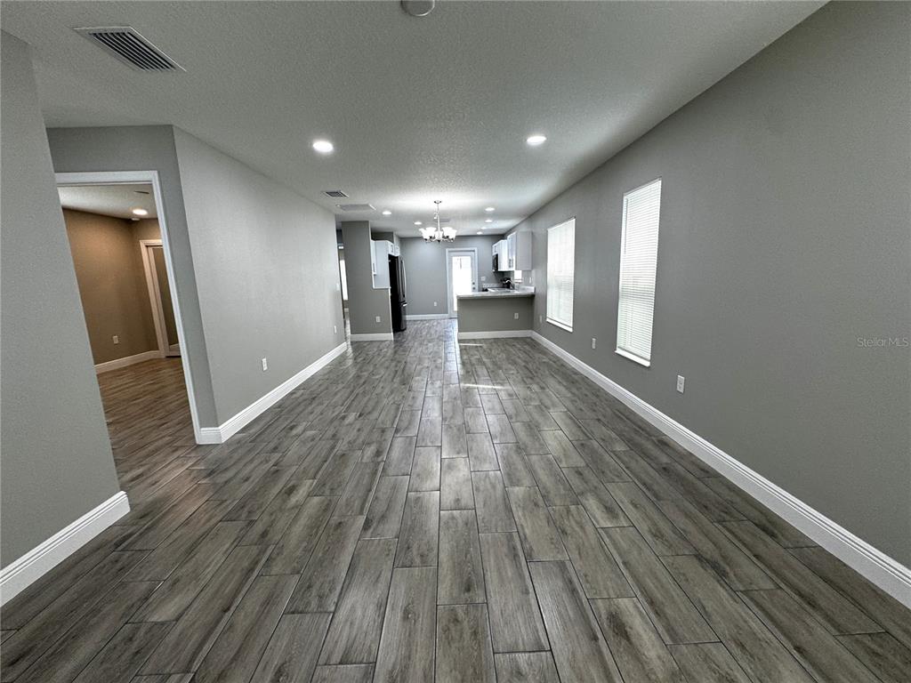 A long, open-concept room features gray walls, wood-look tile flooring, and a distant kitchen area — 5 OLIVE PLACE, OCALA, FL