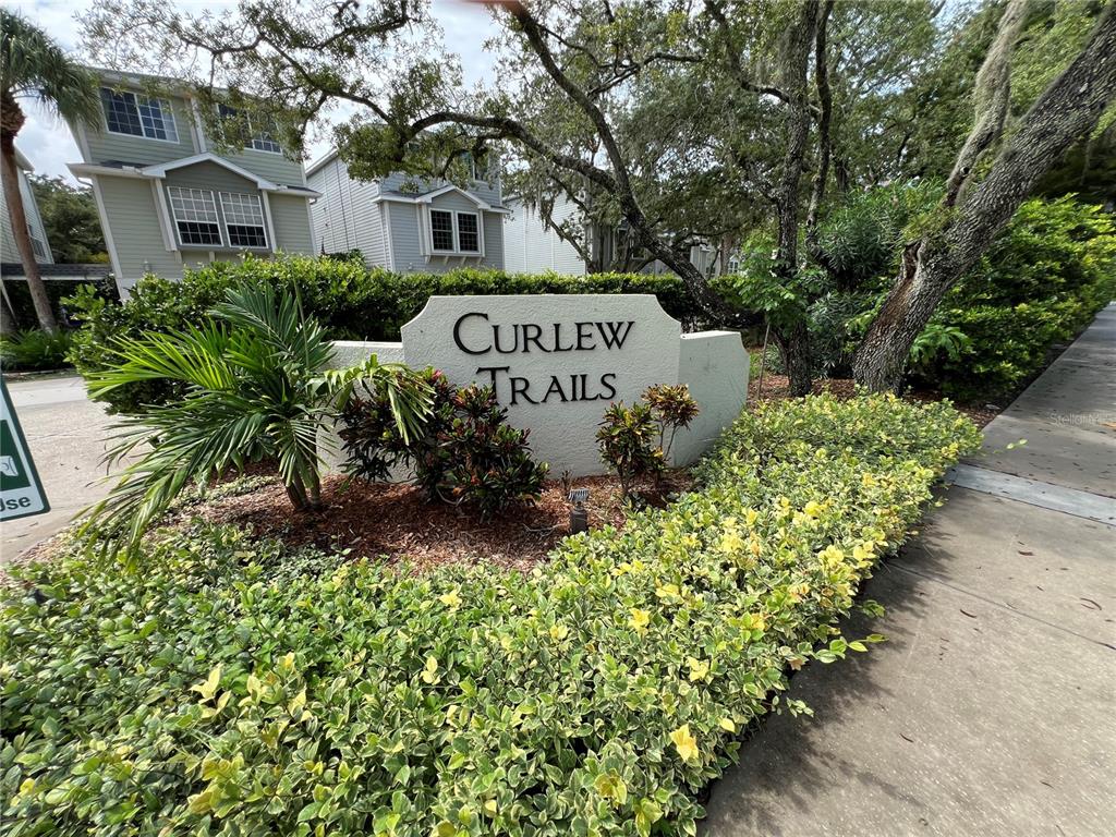A beige stone monument sign for Curlew Trails sits in a landscaped garden area — 1383 CURLEW ROAD, DUNEDIN, FL