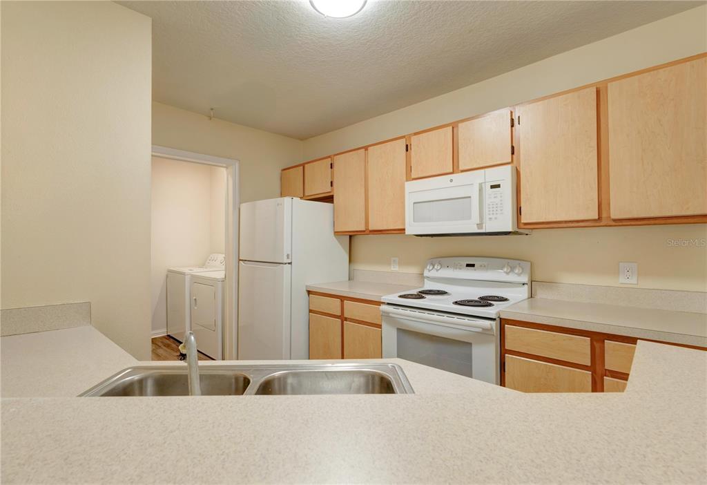 A kitchen features light wood cabinets, white appliances, and laminate countertops overlooking a laundry room — 6566 SWISSCO DRIVE UNIT 425, ORLANDO, FL
