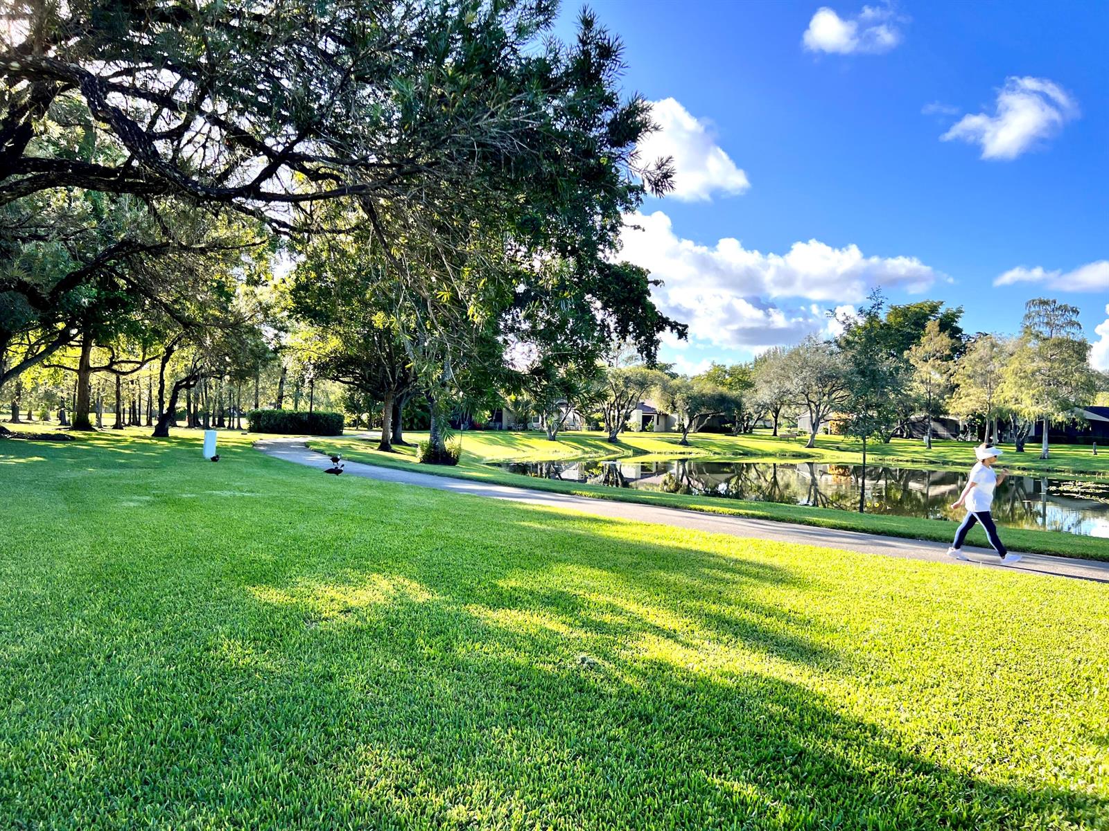 A person walks along a paved path beside a pond in a grassy park area — 22303 Misty Woods Way, Boca Raton FL 33428, BOCA RATON, FL