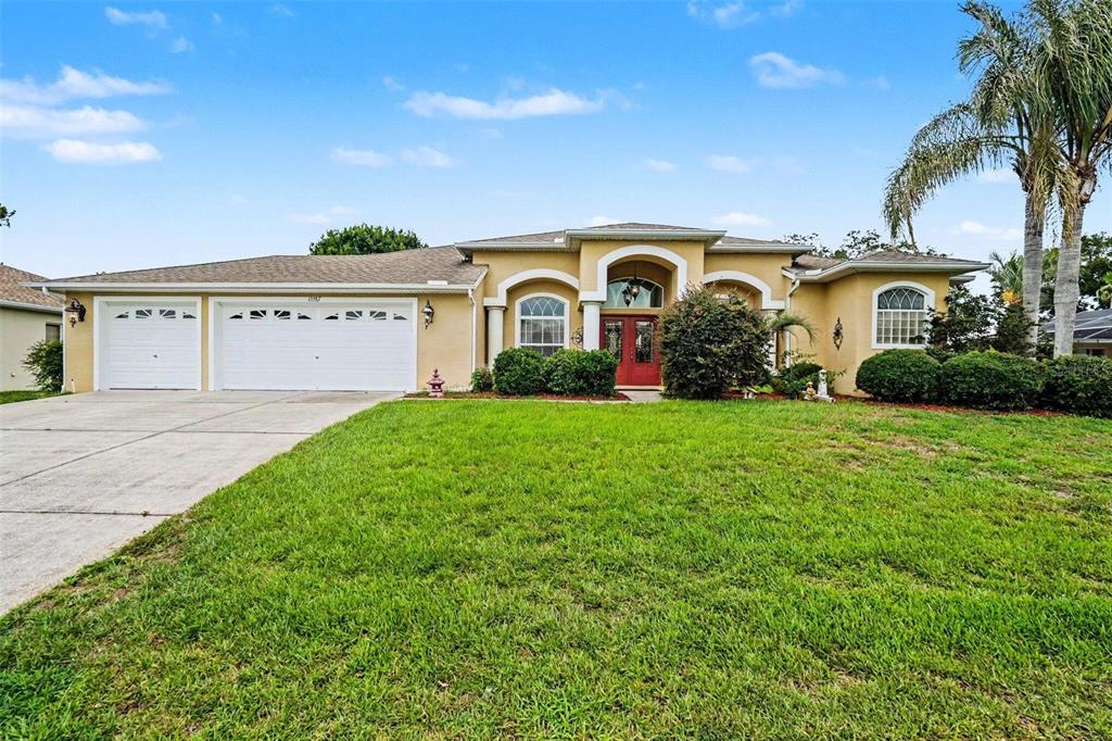 A single-story tan house with a three-car garage and a red front door — 13387 NEWCASTLE AVENUE, SPRING HILL, FL