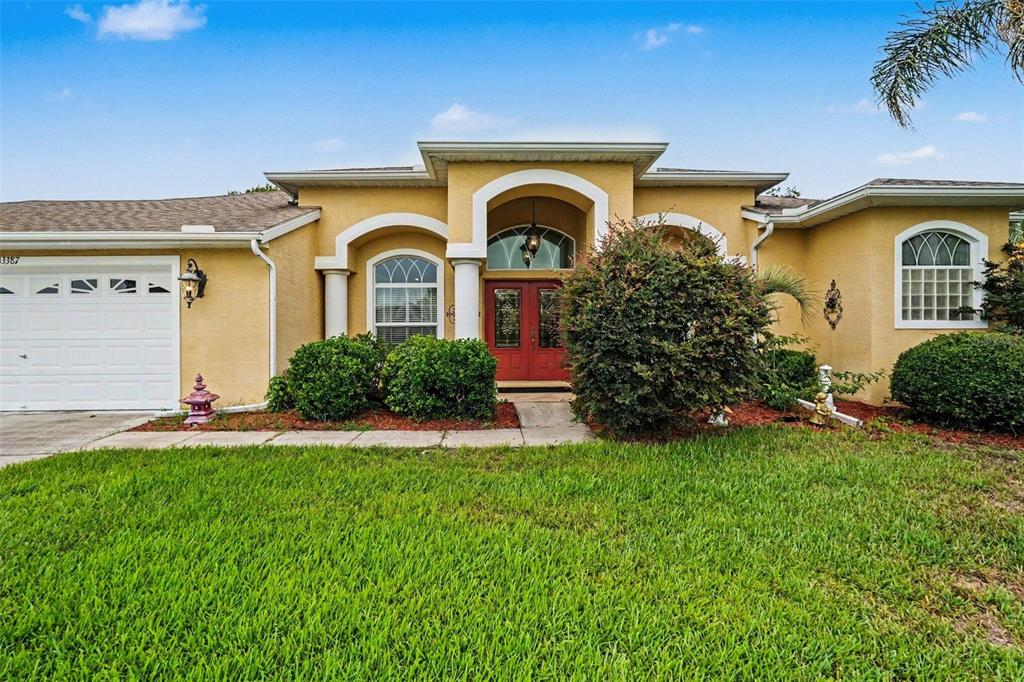 A single-story house with a yellow stucco exterior, red double front doors, and a garage — 13387 NEWCASTLE AVENUE, SPRING HILL, FL