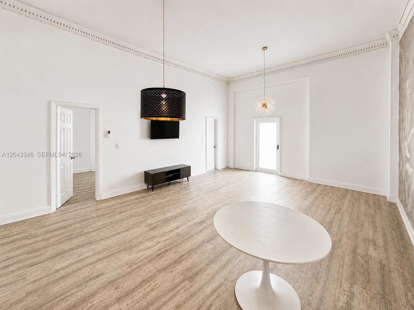 A spacious, high-ceilinged room features light wood-look flooring, two pendant lights, and white walls — 3025 Indian Creek Dr # 110, Miami Beach FL 33140, MIAMI BEACH, FL