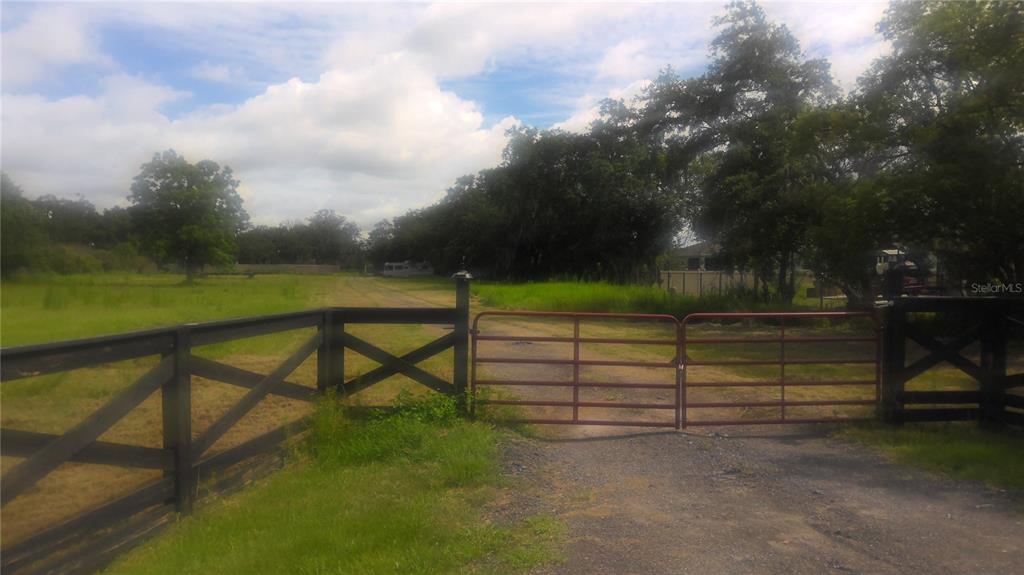 A red metal gate opens onto a gravel driveway leading into a grassy, tree-lined property — 2875 SHARP ROAD, KISSIMMEE, FL