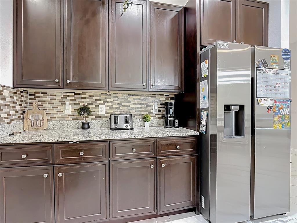 A kitchen features dark wood cabinets, granite countertops, a tiled backsplash, and a stainless refrigerator — 471 GANNET COURT, POINCIANA, FL