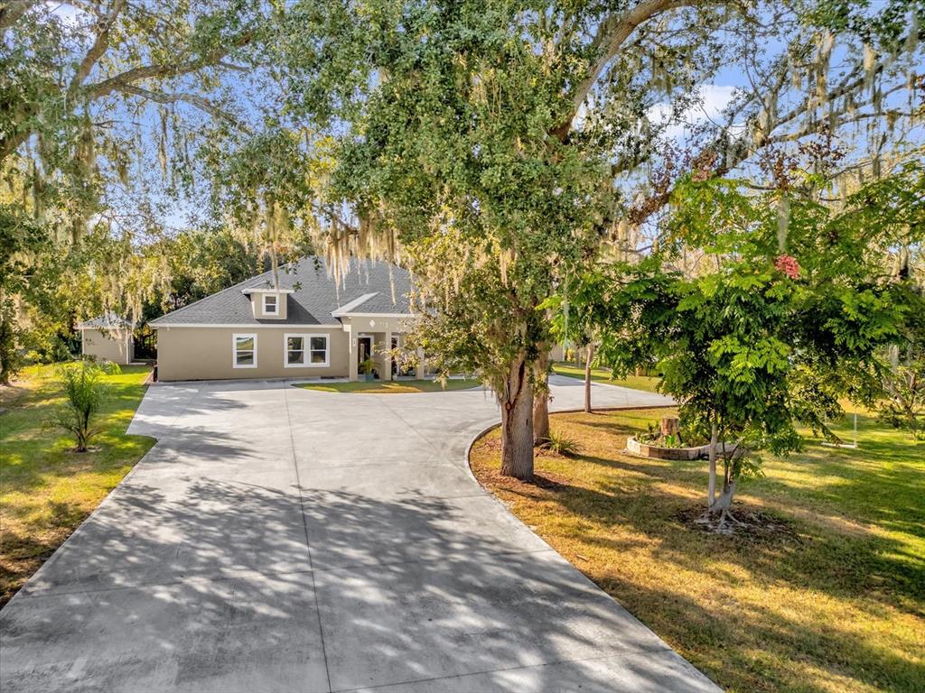 A tan single-story house with a large concrete driveway sits under mature oak trees — 705 LENOX PLACE, KISSIMMEE, FL
