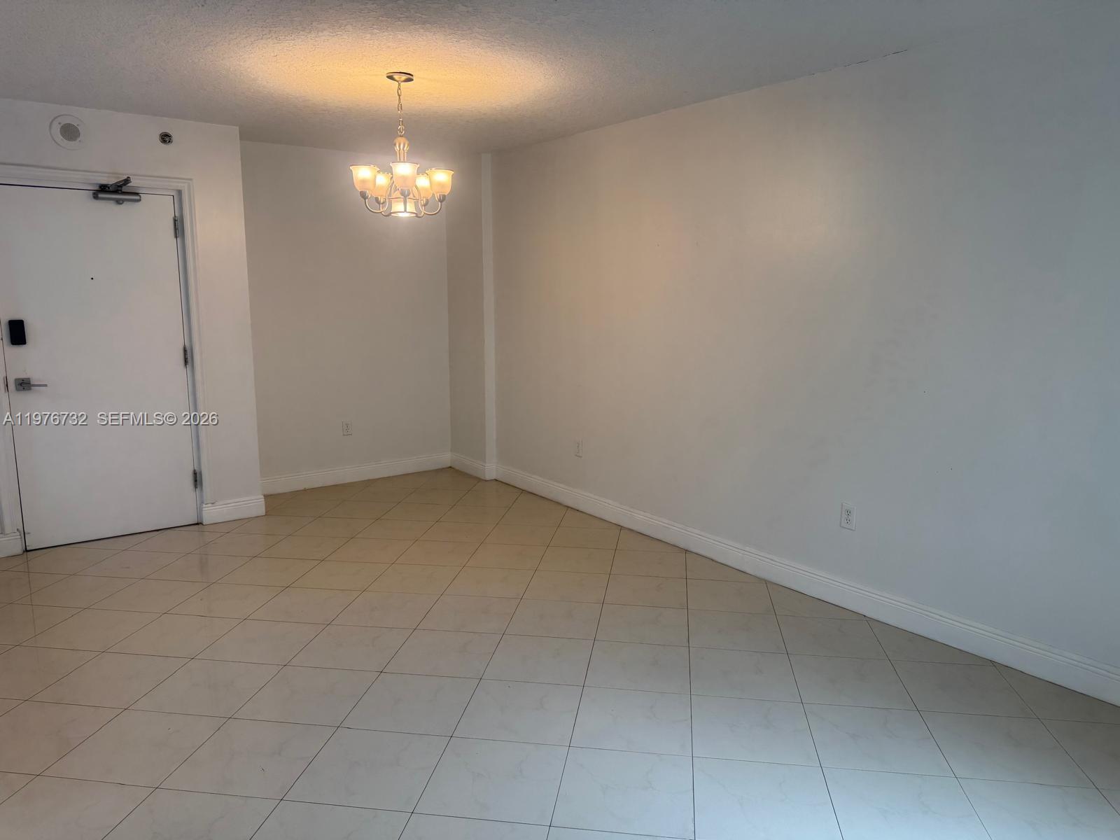 An empty room features white walls, a chandelier, and light-colored tile flooring laid diagonally — 104 SW 9th St # 1403, Miami FL 33130, MIAMI, FL