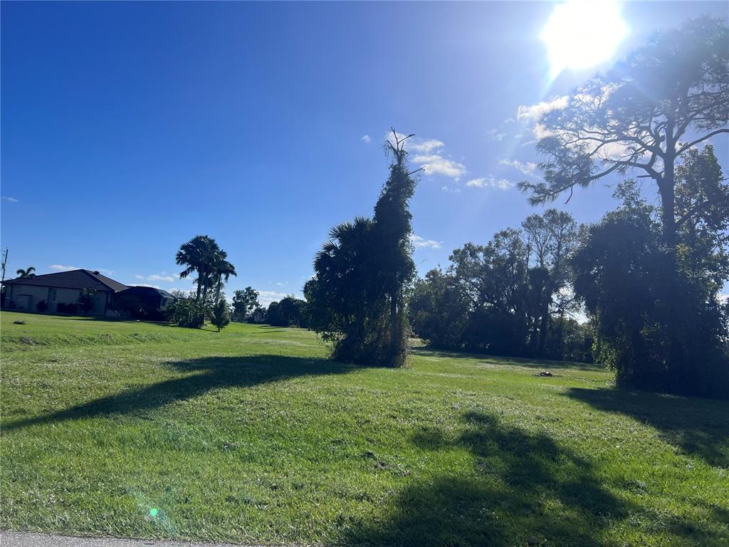 A grassy, sunlit vacant lot features scattered trees and a house in the distance — 16818 ACAPULCO ROAD, PUNTA GORDA, FL