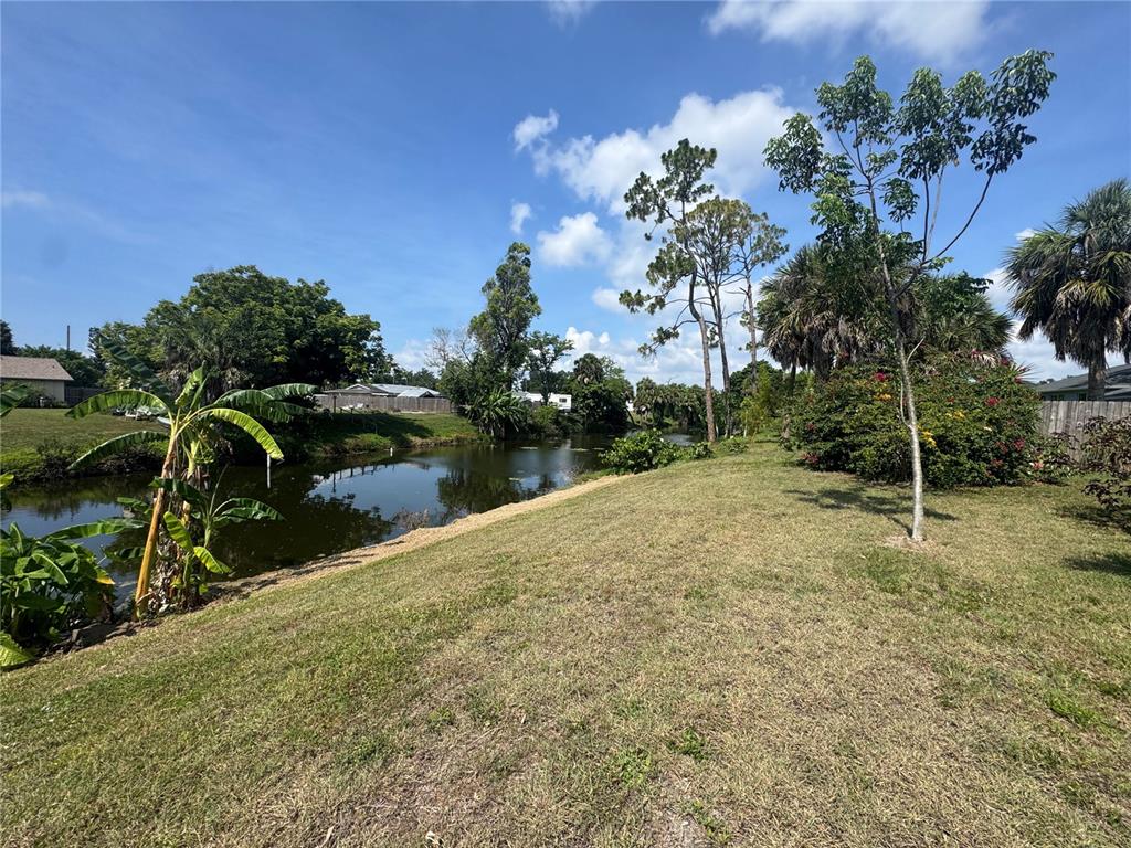 A grassy backyard slopes down toward a canal lined with trees and tropical plants — 21239 COTTONWOOD AVENUE, PORT CHARLOTTE, FL