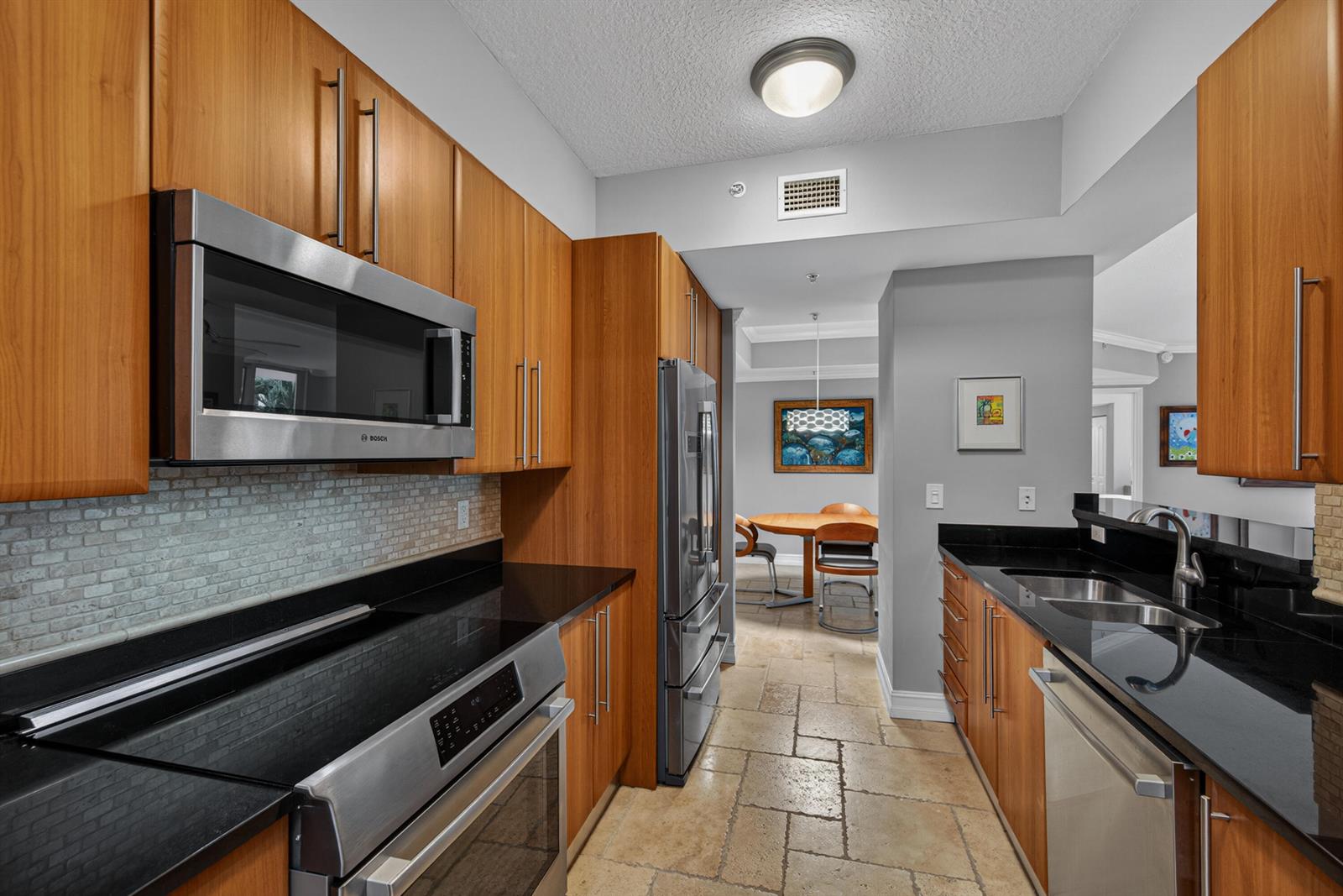 A galley kitchen features wood cabinets, black countertops, stainless steel appliances, and stone tile flooring — 101 SE 20th Avenue # 201, Deerfield Beach FL 33441, DEERFIELD BEACH, FL