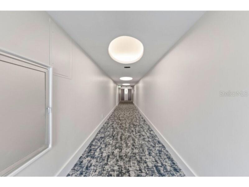 A long, narrow indoor hallway features white walls, recessed circular ceiling lights, and patterned carpet — 1111 N GULFSTREAM AVENUE UNIT 5B, SARASOTA, FL