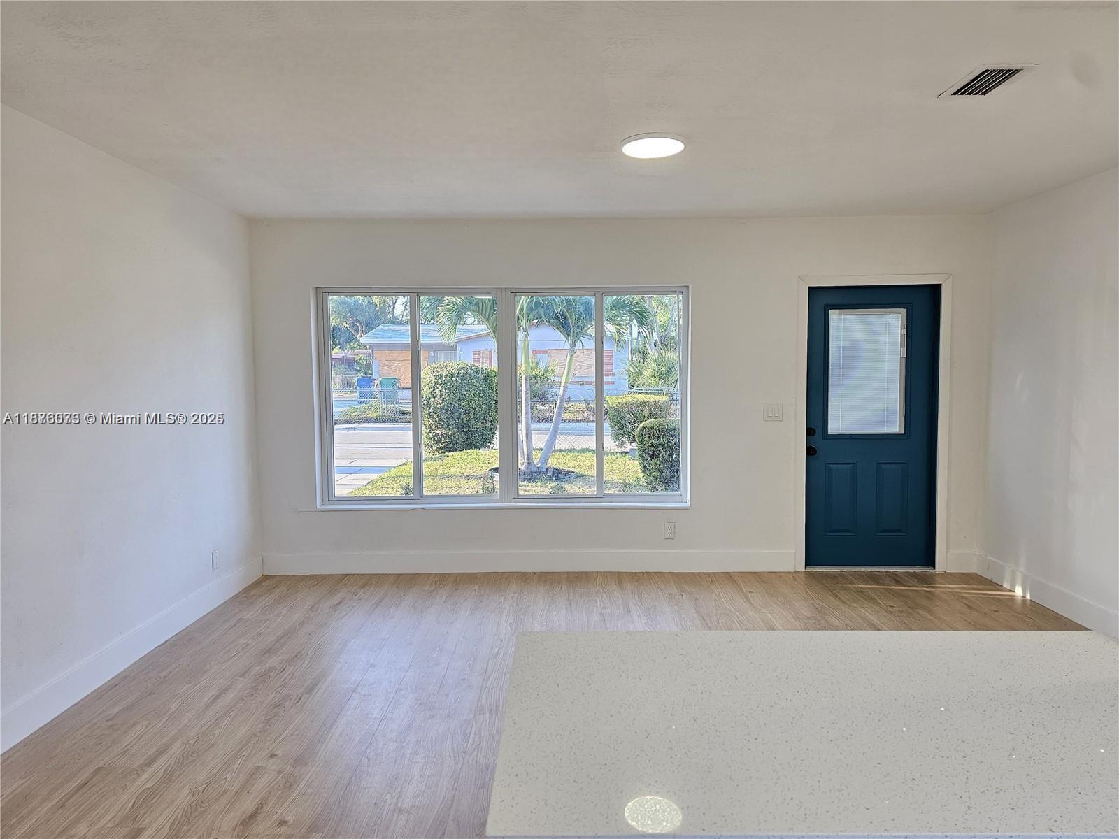 A bright room features light wood-look flooring, white walls, a large window, and a blue door — 1117 W 26th Ct, Riviera Beach FL 33404, RIVIERA BEACH, FL