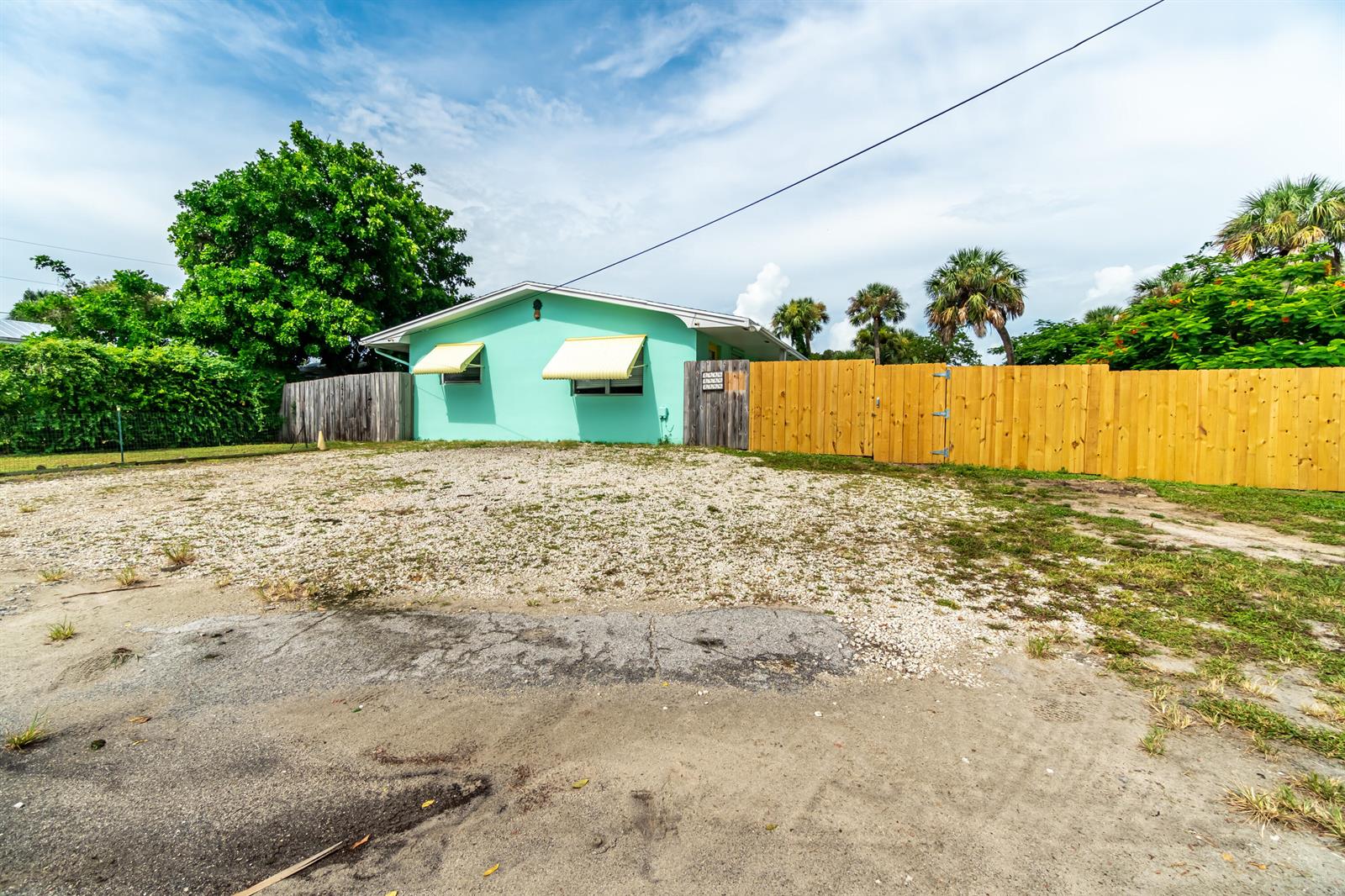 A light green house with yellow awnings sits behind a wooden fence and gravel yard — 2546 NE Hickory Avenue, Jensen Beach FL 34957, JENSEN BEACH, FL
