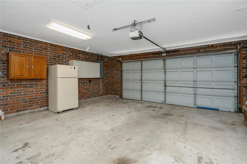 A two-car garage features a brick wall, a refrigerator, storage cabinets, and a concrete floor — 785 DUQUE ROAD, LUTZ, FL