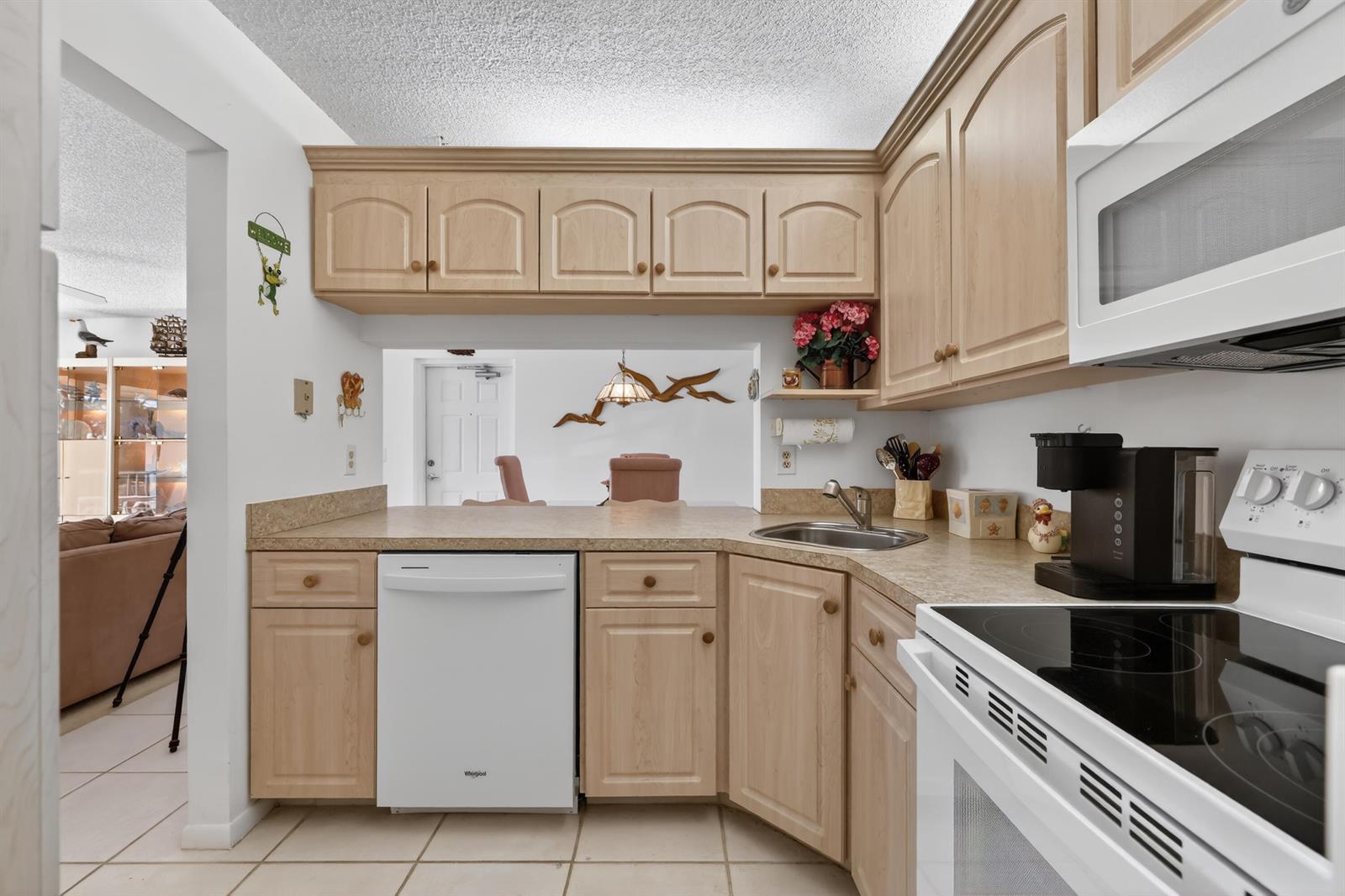 A kitchen features light wood cabinets, white appliances, and a pass-through window to the dining area — 4444 NE Ocean Boulevard # 108, Jensen Beach FL 34957, JENSEN BEACH, FL