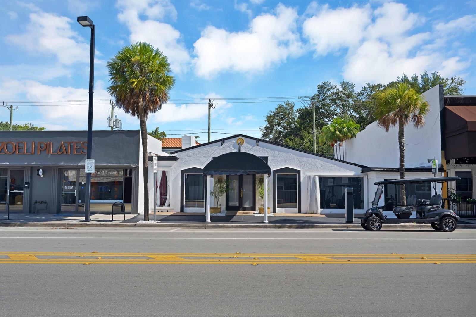 A white commercial building with a black awning stands along a street with palm trees — FORT LAUDERDALE, FL