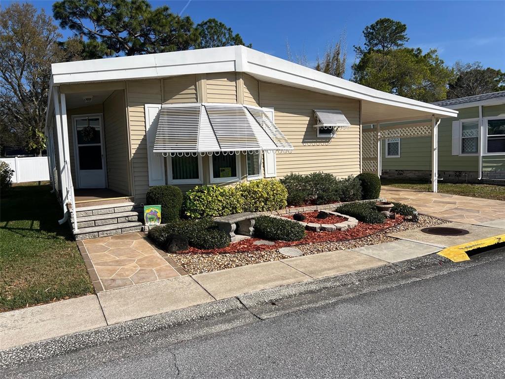 A beige mobile home with a front porch, carport, and landscaped yard under a blue sky — 1001 STARKEY ROAD UNIT 90, LARGO, FL