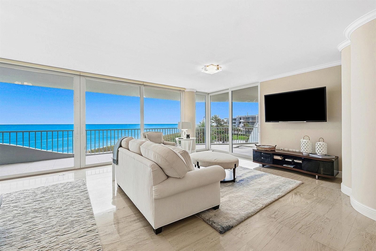 A living room features a beige sofa, a television, and floor-to-ceiling windows overlooking the ocean — 2001 SE Sailfish Point Boulevard # 313, Stuart FL 34996, STUART, FL