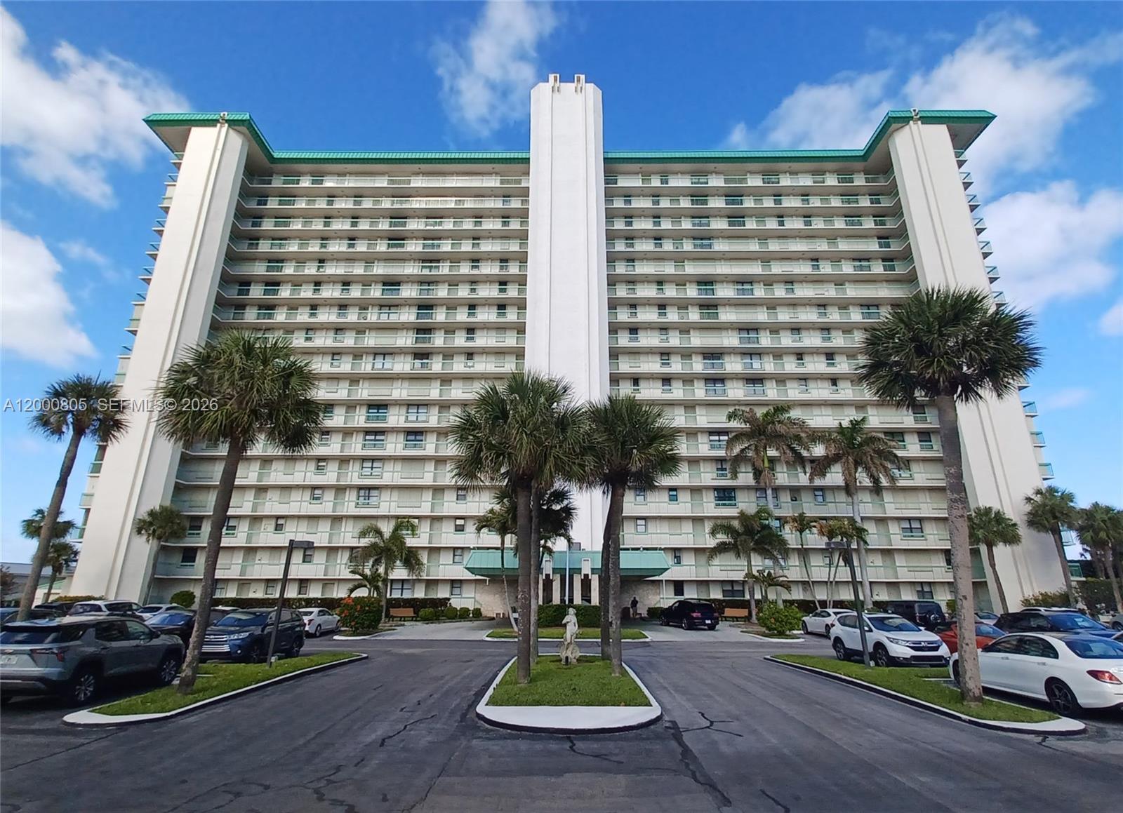 A tall, multi-story residential building with balconies stands behind a paved parking lot and palm trees — 9900 S Ocean Dr # 106, Jensen Beach FL 34957, JENSEN BEACH, FL