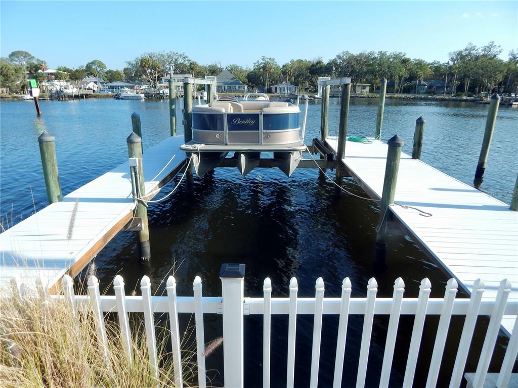 A pontoon boat is lifted on a dock in a canal behind a white fence — 8025 ISLAND DRIVE, PORT RICHEY, FL