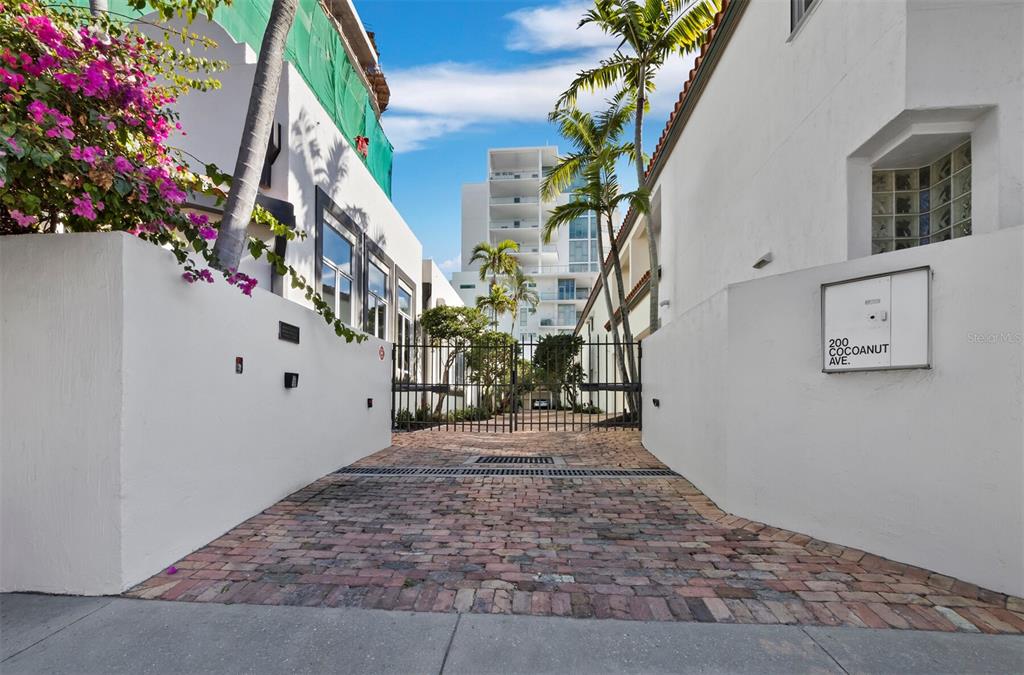 A brick driveway leads to a gated entrance between two white buildings with palm trees — 200 COCOANUT AVENUE UNIT 7, SARASOTA, FL