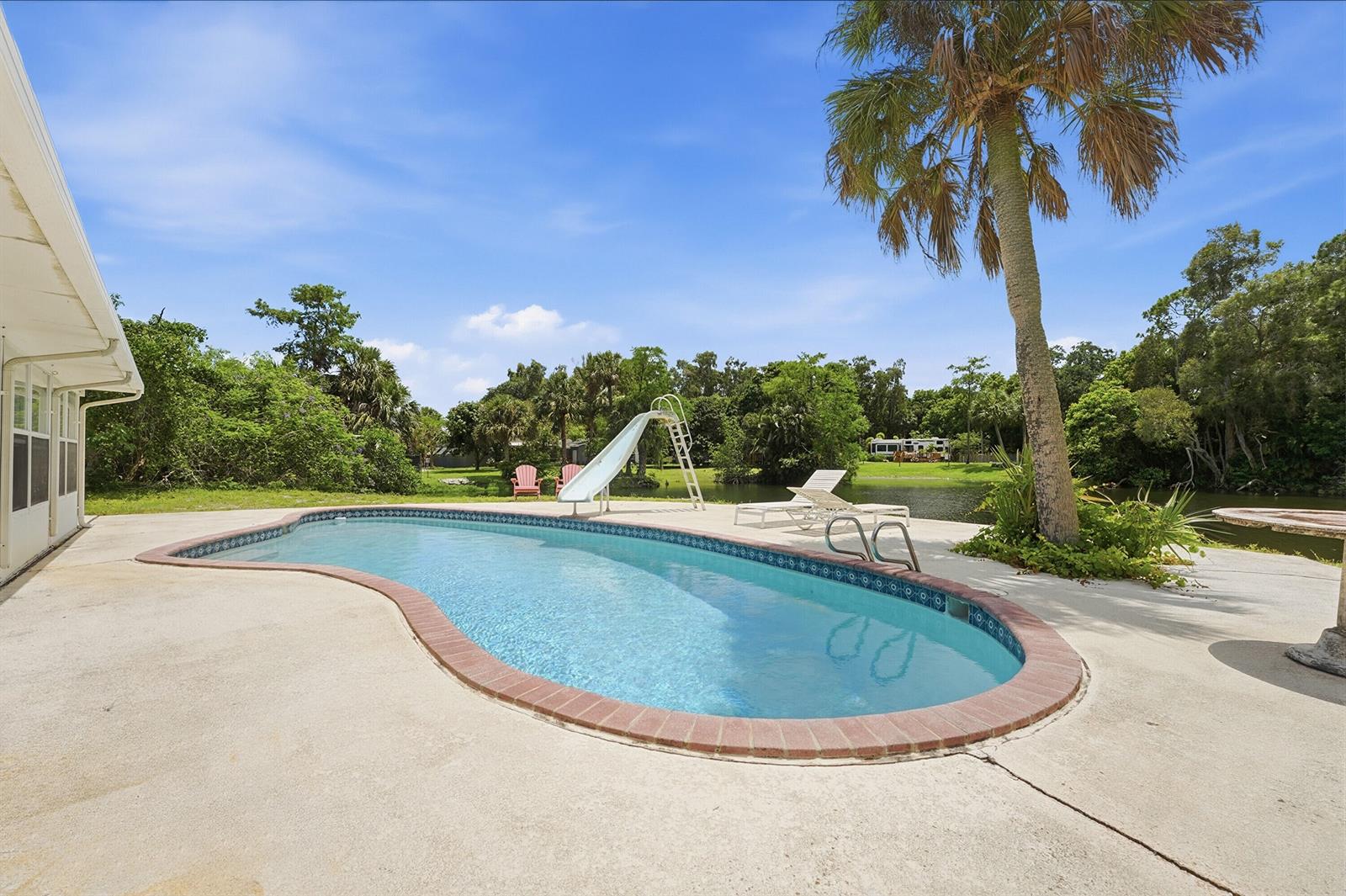 A kidney-shaped swimming pool with a slide sits on a patio overlooking a pond — 7689 Lawrence Road, Boynton Beach FL 33436, BOYNTON BEACH, FL