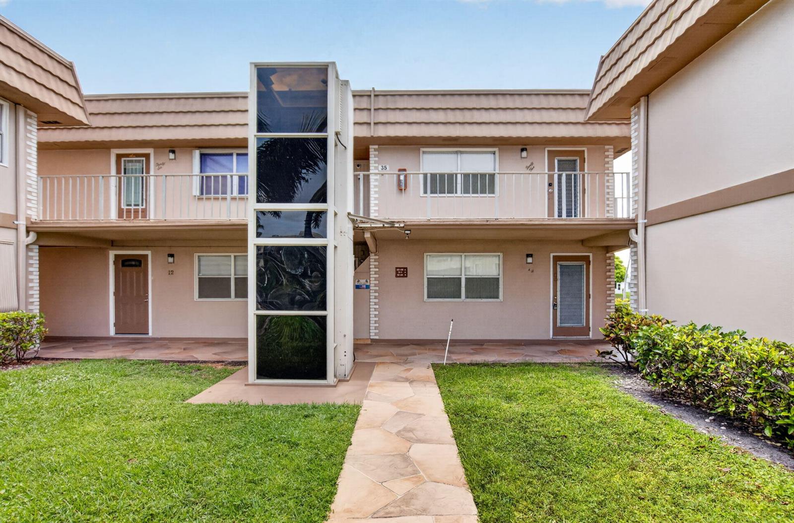 A two-story apartment building exterior features a central glass elevator shaft and stone walkway — 11 Brittany A, Delray Beach FL 33446, DELRAY BEACH, FL
