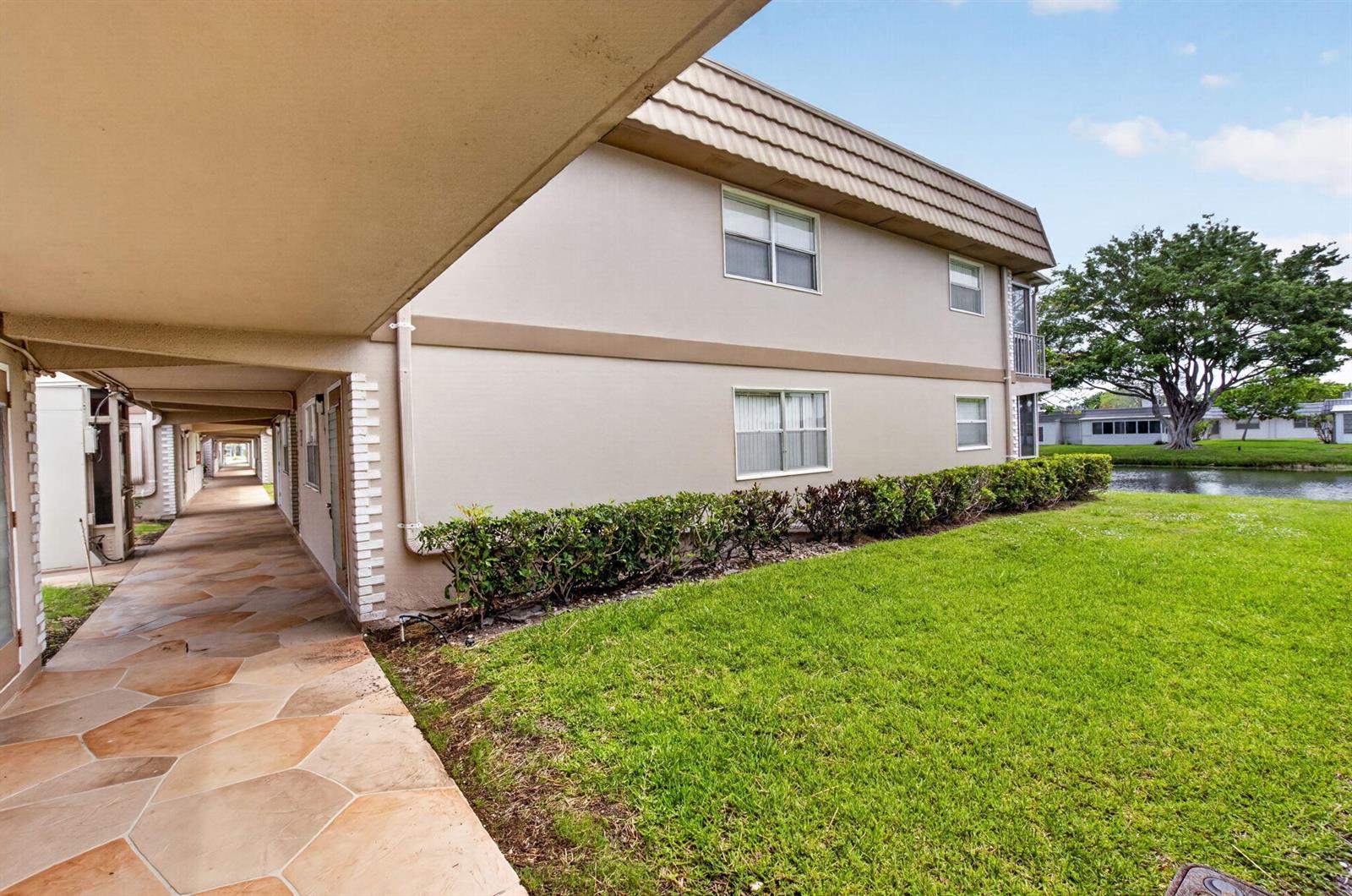 A covered walkway with stone pavers runs alongside a two-story apartment building near a pond — 11 Brittany A, Delray Beach FL 33446, DELRAY BEACH, FL