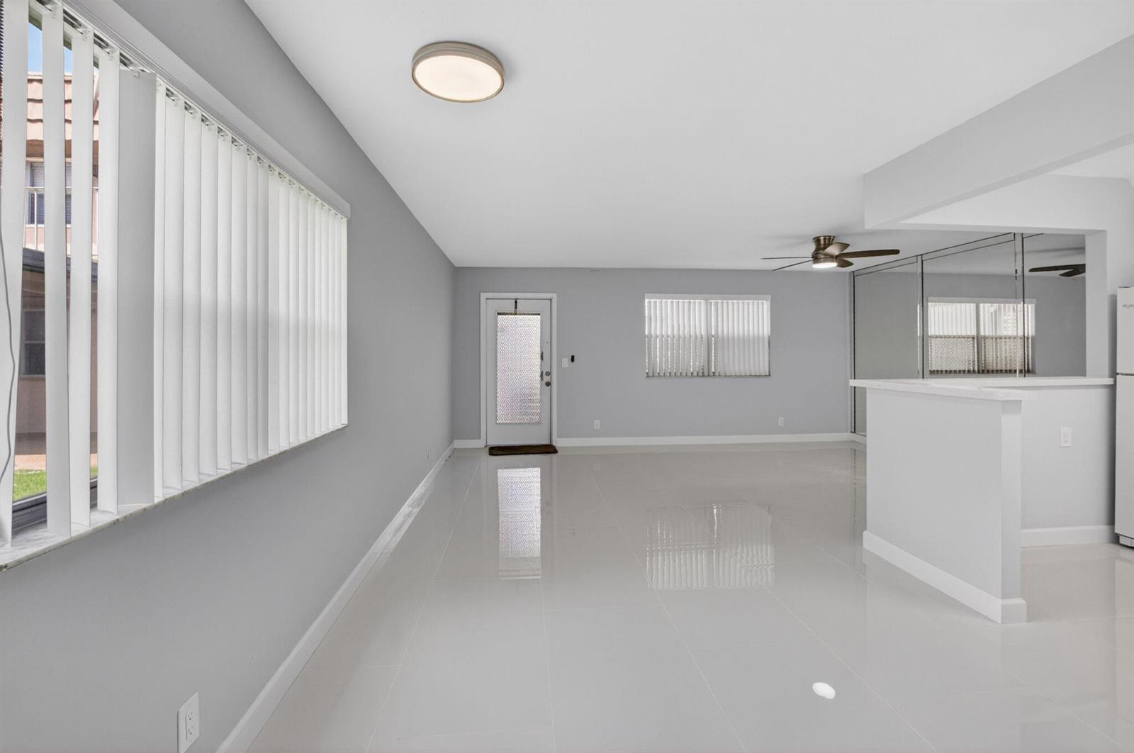 A bright, empty living room with white tile flooring, gray walls, and vertical window blinds — 11 Brittany A, Delray Beach FL 33446, DELRAY BEACH, FL