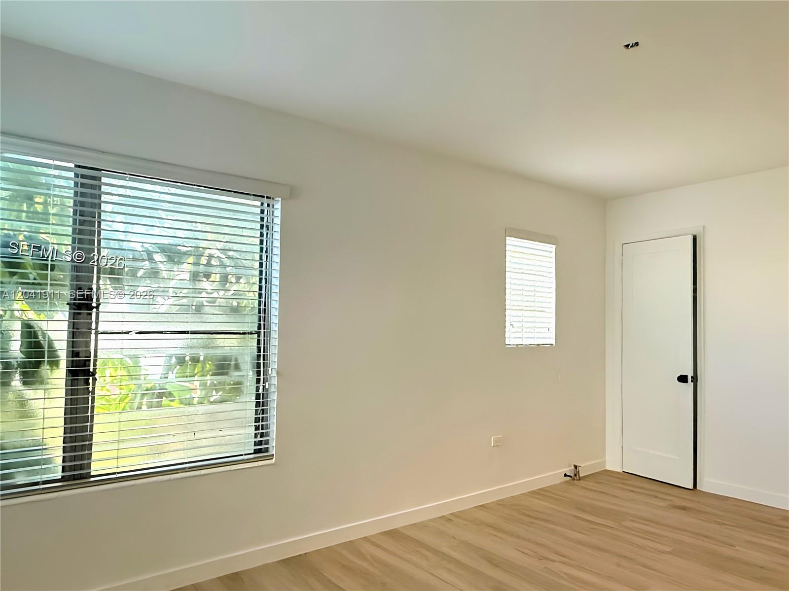 An empty room features white walls, light wood-look flooring, two windows with blinds, and a door — 641 10th St # 2, Miami Beach FL 33139, MIAMI BEACH, FL