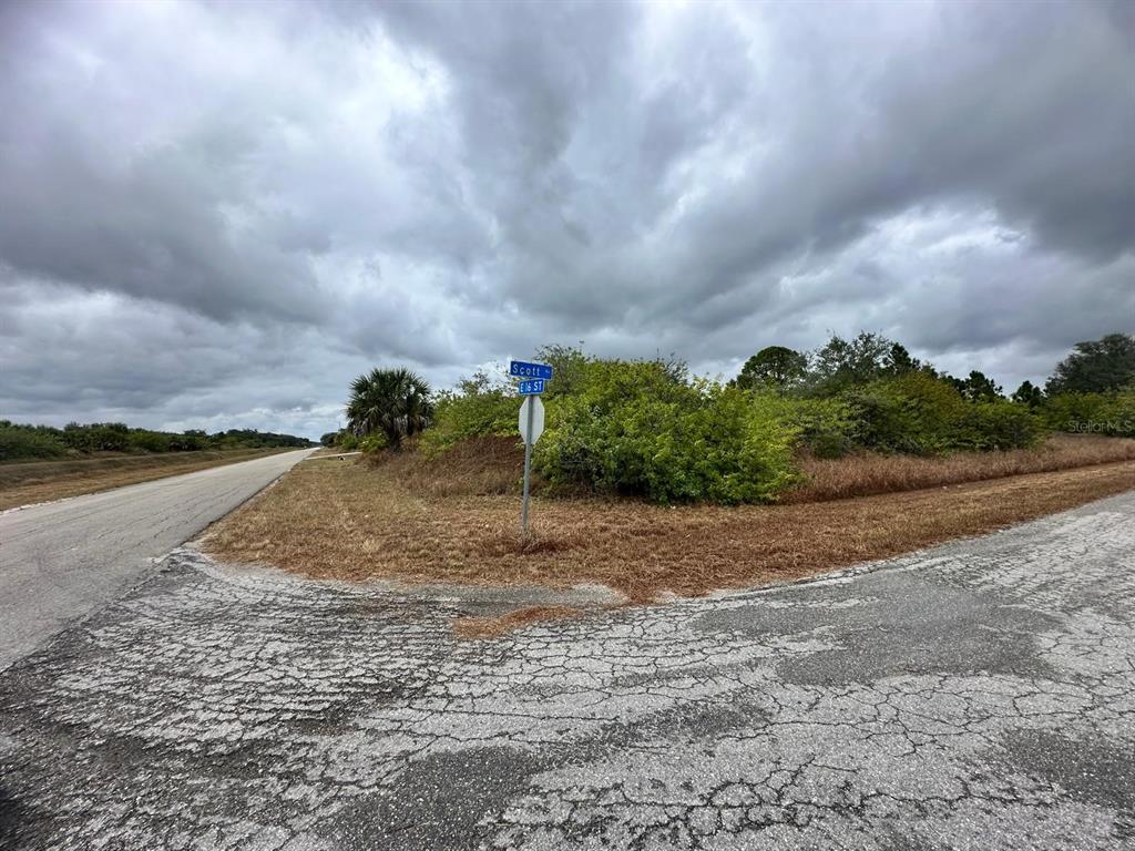A street sign stands at a corner intersection next to a vacant, brush-covered lot — 1610 SCOTT AVENUE, LEHIGH ACRES, FL