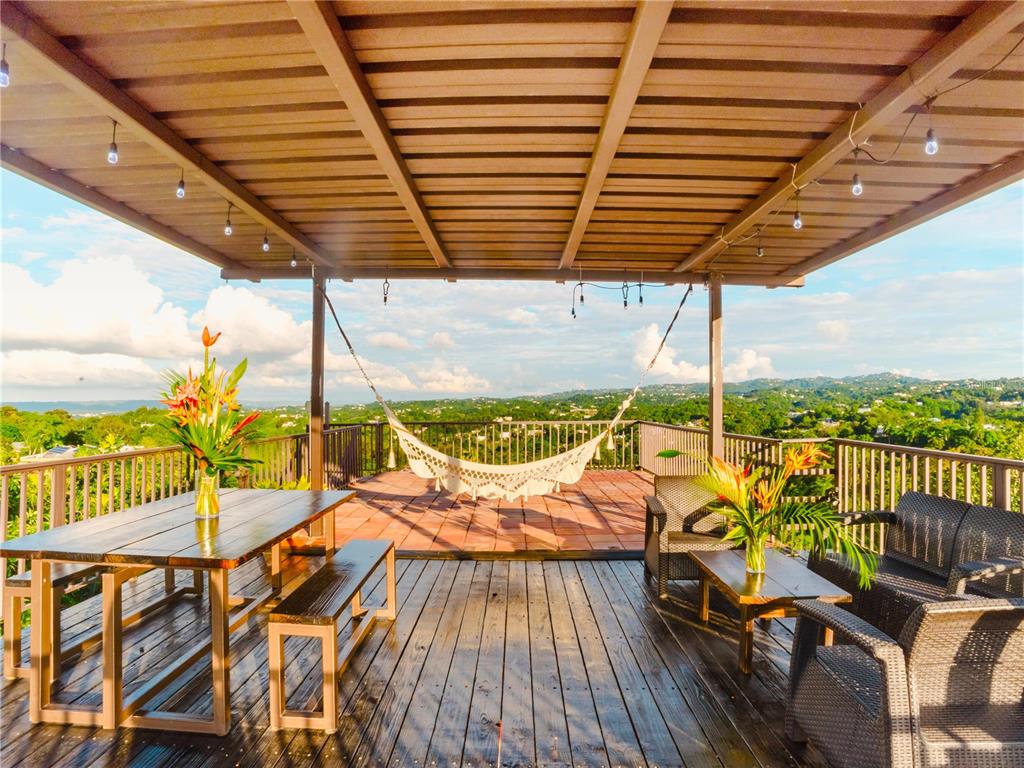 A covered wooden deck features a dining set, lounge furniture, and a hammock overlooking hills — CARR. 414 KM 1.8 INT THE TERRACES, RINCON, PR