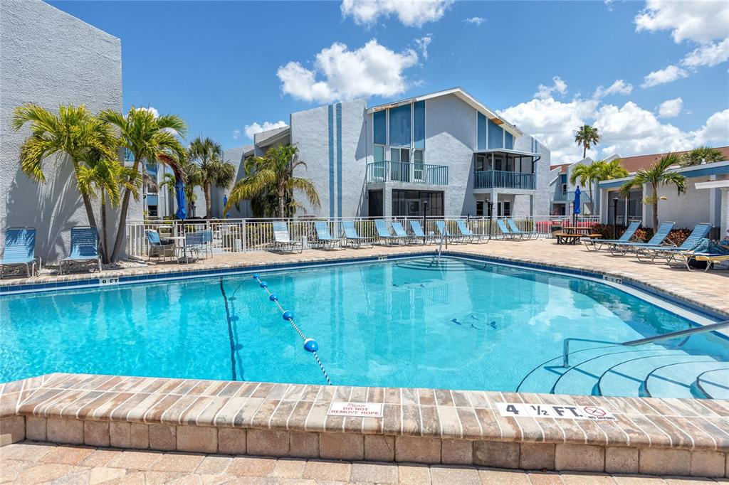 A swimming pool with lounge chairs sits in front of a multi-story apartment building — 159 MEDALLION BOULEVARD UNIT F, MADEIRA BEACH, FL