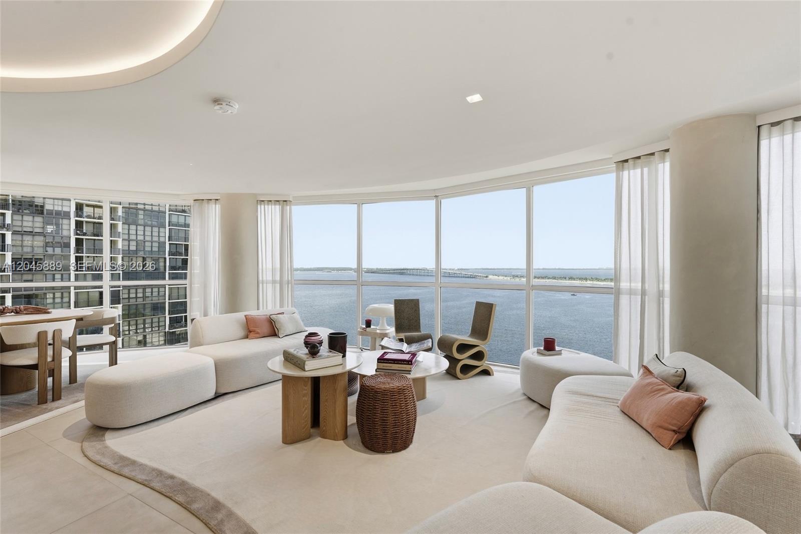 A modern living room features curved sofas, a round coffee table, and floor-to-ceiling windows overlooking water — 2025 Brickell Ave # 1401, Miami FL 33129, MIAMI, FL