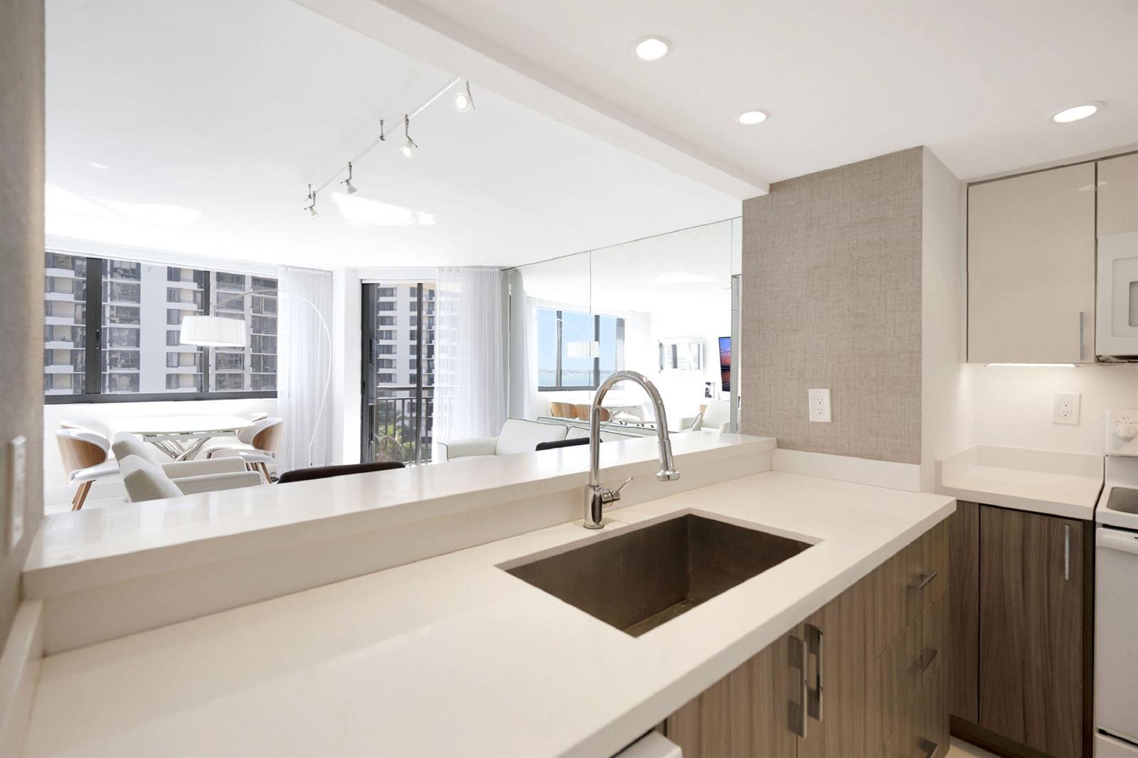 A modern kitchen with white countertops and wood cabinets overlooks a bright living area — 540 Brickell Key Drive # 919, Miami FL 33131, MIAMI, FL