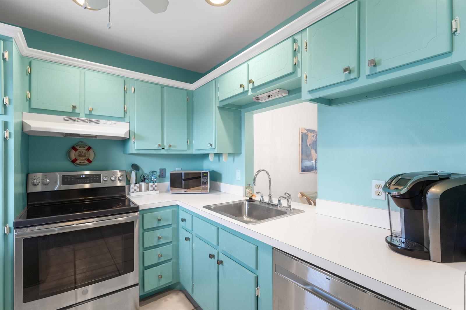 A kitchen features turquoise cabinets, white countertops, stainless steel appliances, and a pass-through window — 3851 N Ocean Boulevard Unit 404, Delray Beach FL 33483, DELRAY BEACH, FL