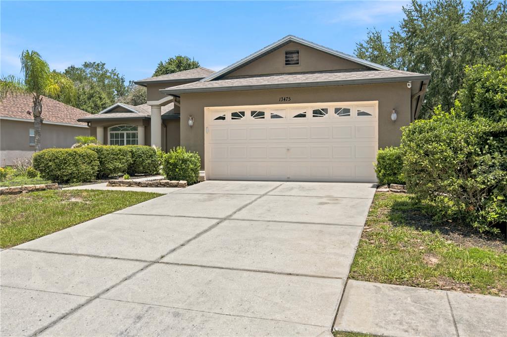 A tan single-story house features a two-car garage, a concrete driveway, and landscaped front bushes — 13475 TEABERRY LANE, SPRING HILL, FL
