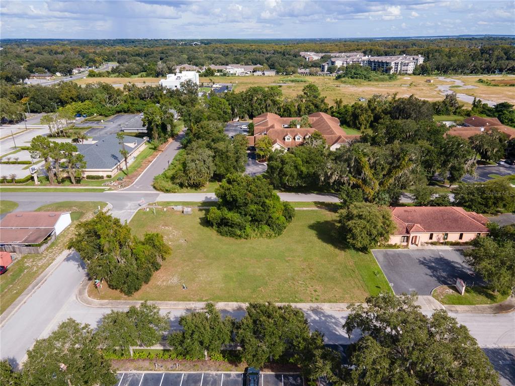 An aerial view shows a vacant grassy lot surrounded by trees, parking lots, and buildings — 700 PHYSICIANS COURT, LEESBURG, FL