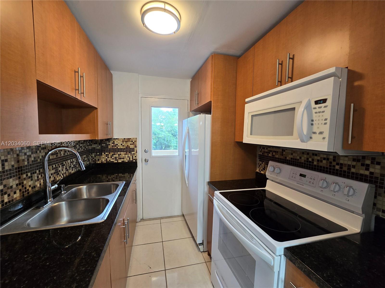 A galley kitchen features wood cabinets, black countertops, a mosaic backsplash, and white appliances — 1475 NE 125th Ter # B, North Miami FL 33161, NORTH MIAMI, FL