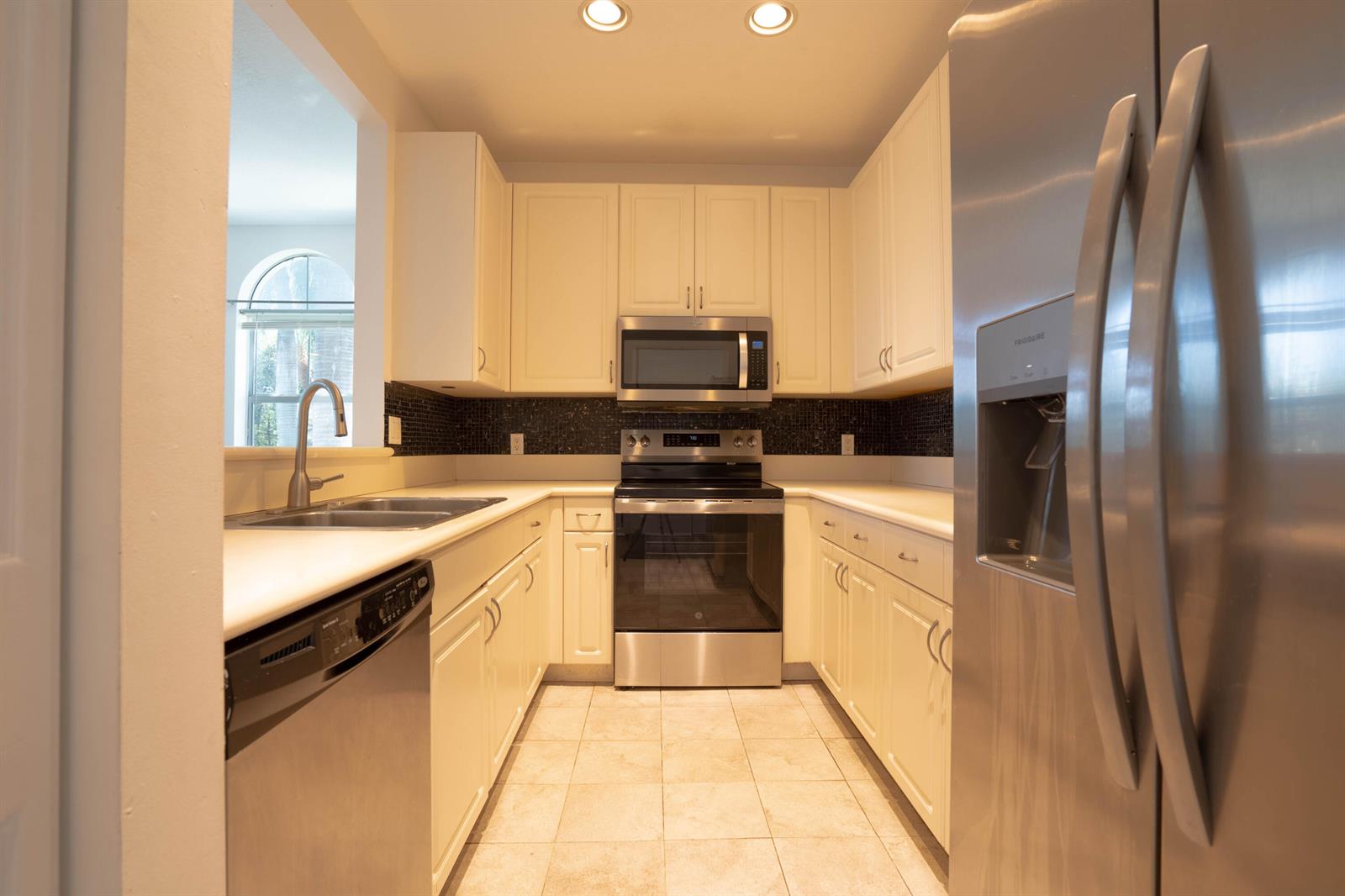 A galley kitchen features white cabinets, stainless steel appliances, and a dark tiled backsplash — 1356 Piazza Pitti, Boynton Beach FL 33426, BOYNTON BEACH, FL