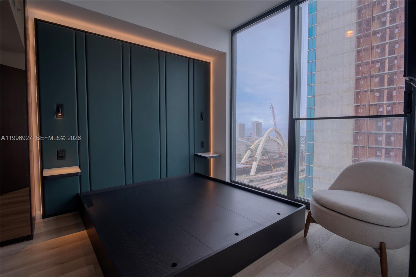 A bedroom features a dark platform bed, teal paneled wall, and floor-to-ceiling windows overlooking construction — 20 NE 11th st # 2308, Miami FL 33132, MIAMI, FL
