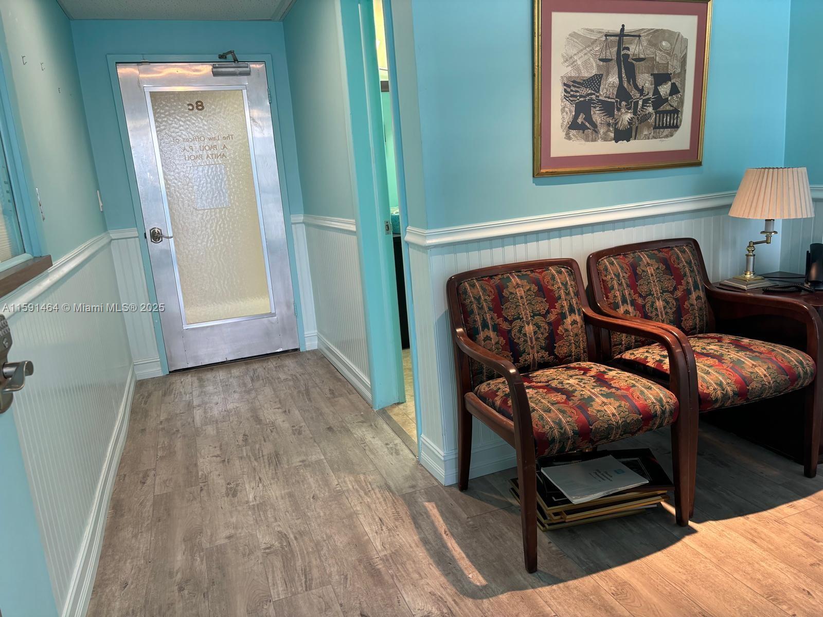 A waiting area features light blue walls, wood-look flooring, two patterned chairs, and a metal door — 1720 Harrison St # 8C, Hollywood FL 33020, HOLLYWOOD, FL