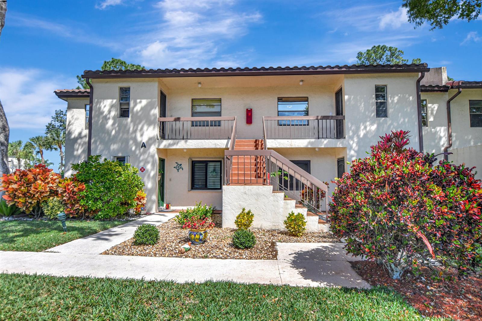 A two-story apartment building exterior features a central staircase, balconies, and landscaped front gardens — 21864 Arriba Real # 3d, Boca Raton FL 33433, BOCA RATON, FL