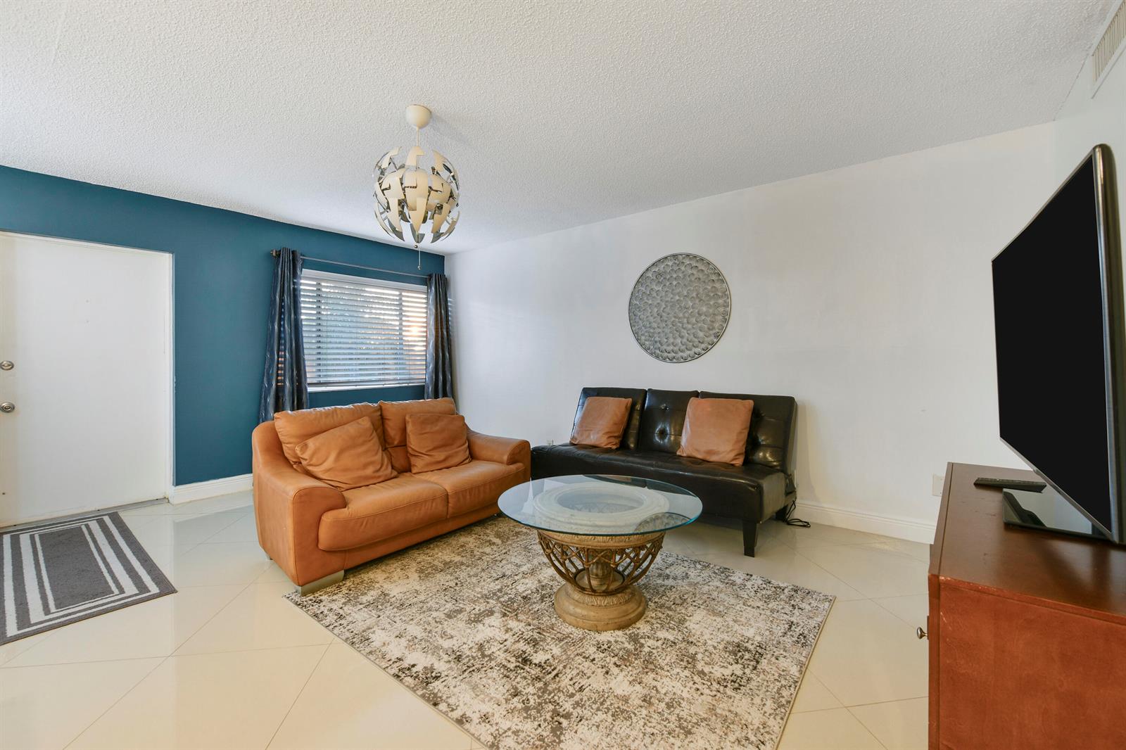 A living room features a tan sofa, a black futon, a glass coffee table, and tile flooring — 3100 Springdale Boulevard # 314, Palm Springs FL 33461, PALM SPRINGS, FL
