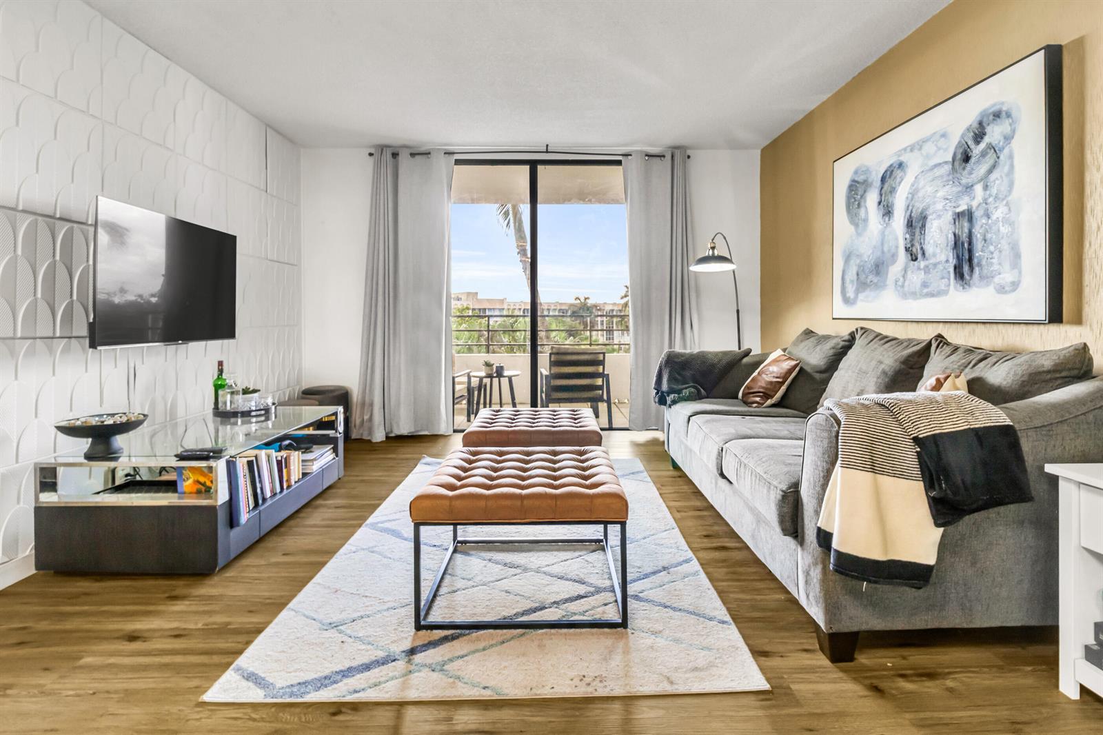 A living room features a grey sofa, two leather ottomans, a glass media console, and balcony access — 500 Three Islands Boulevard # 109, Hallandale Beach FL 33009, HALLANDALE BEACH, FL