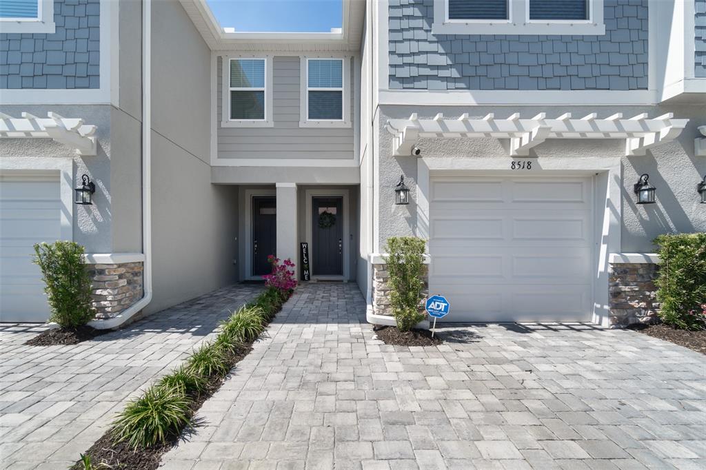 A two-story townhouse exterior features a paver driveway, white garage doors, and gray siding — 8518 PRIMROSE WILLOW PLACE, ODESSA, FL