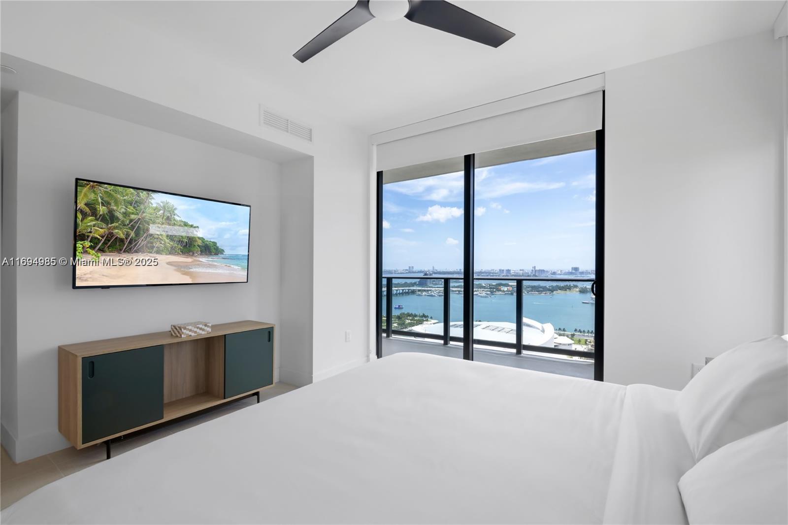 A modern bedroom features a wall-mounted television, a media console, and a balcony with water views — 159 NE 6TH ST # 2916, Miami FL 33132, MIAMI, FL