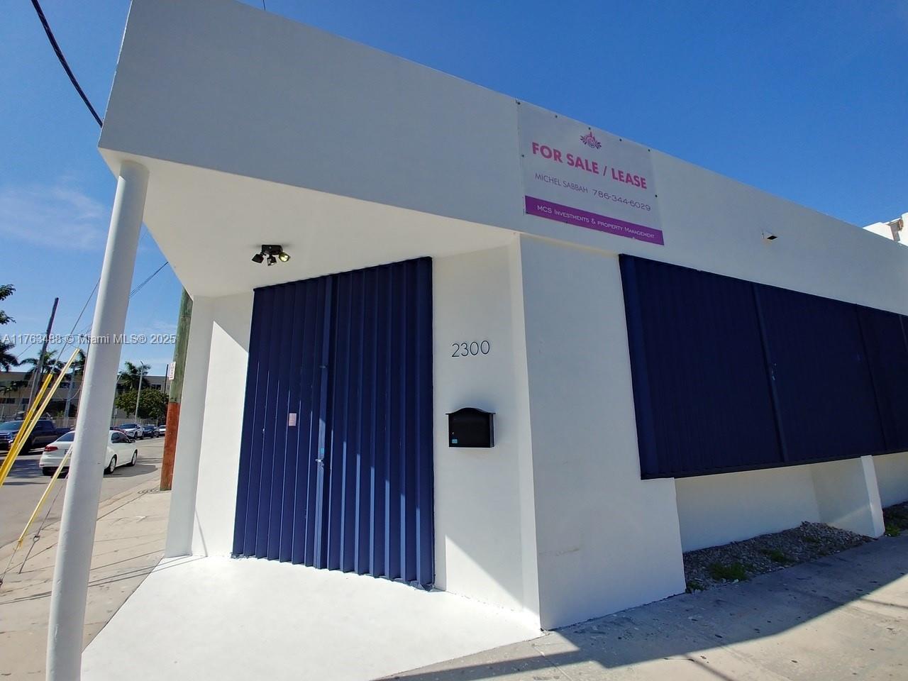 A white commercial building exterior features a blue accordion door and a for sale sign — 2300 N Miami Ave, Miami FL 33127, MIAMI, FL