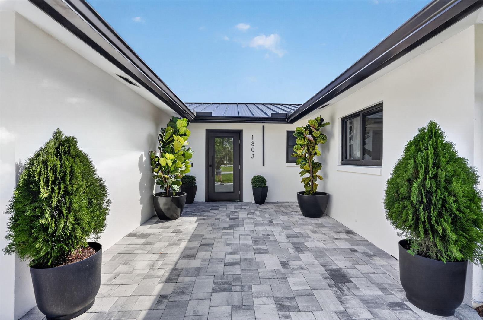 A paved entryway leads to a modern front door flanked by potted plants and trees — 1803 Juno Isles Boulevard, North Palm Beach FL 33408, NORTH PALM BEACH, FL